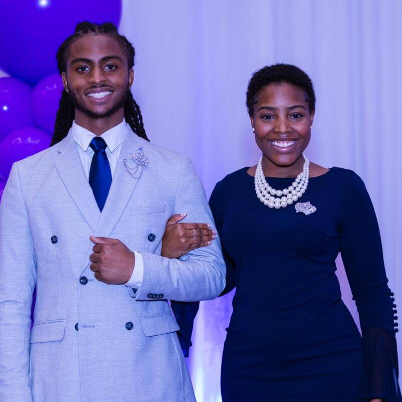Two people posing for photo in front of a lavender colored background